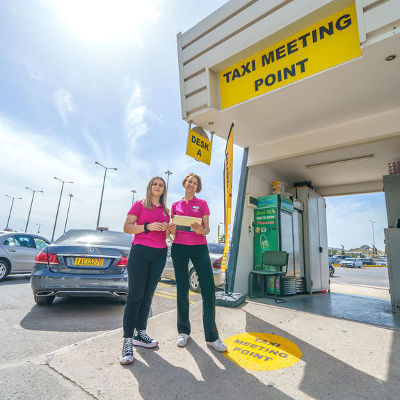 Taxi meeting point at Heraklion Airport with staff ready to assist, clear signage and parked vehicles nearby
