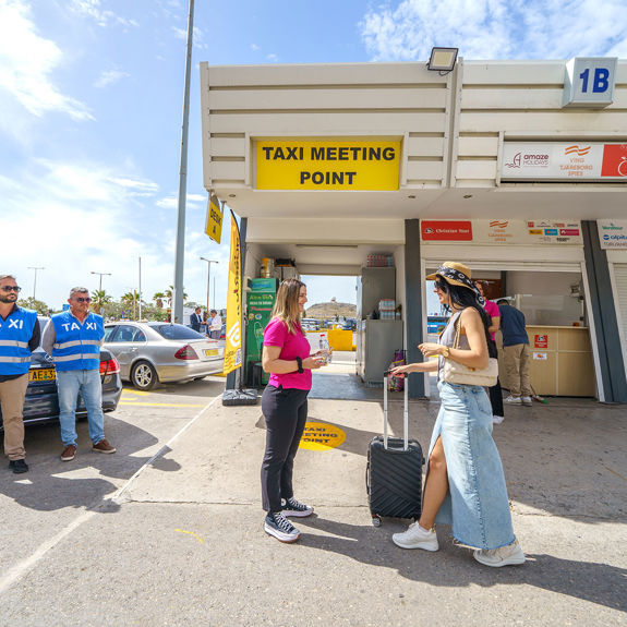 Taxi meeting point at Heraklion Airport with staff arriving passenger with luggage, drivers waiting nearby
