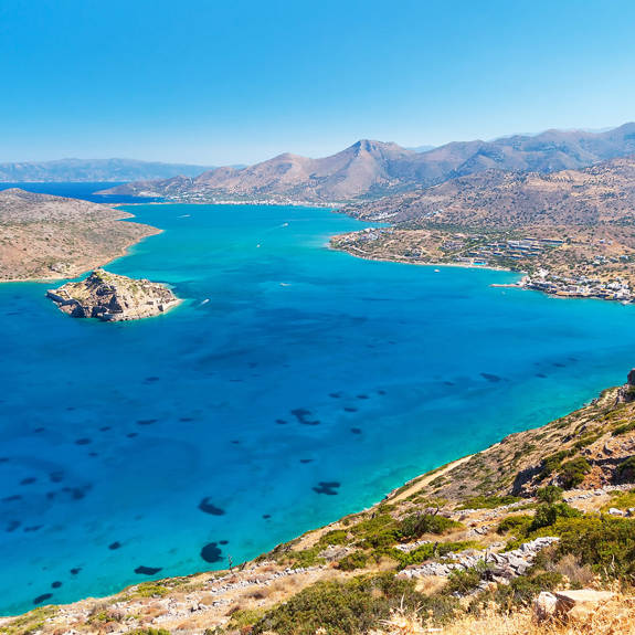 Aerial view of Elounda Crete with blue lagoon, small island, coastal town and surrounding hills under clear sky
